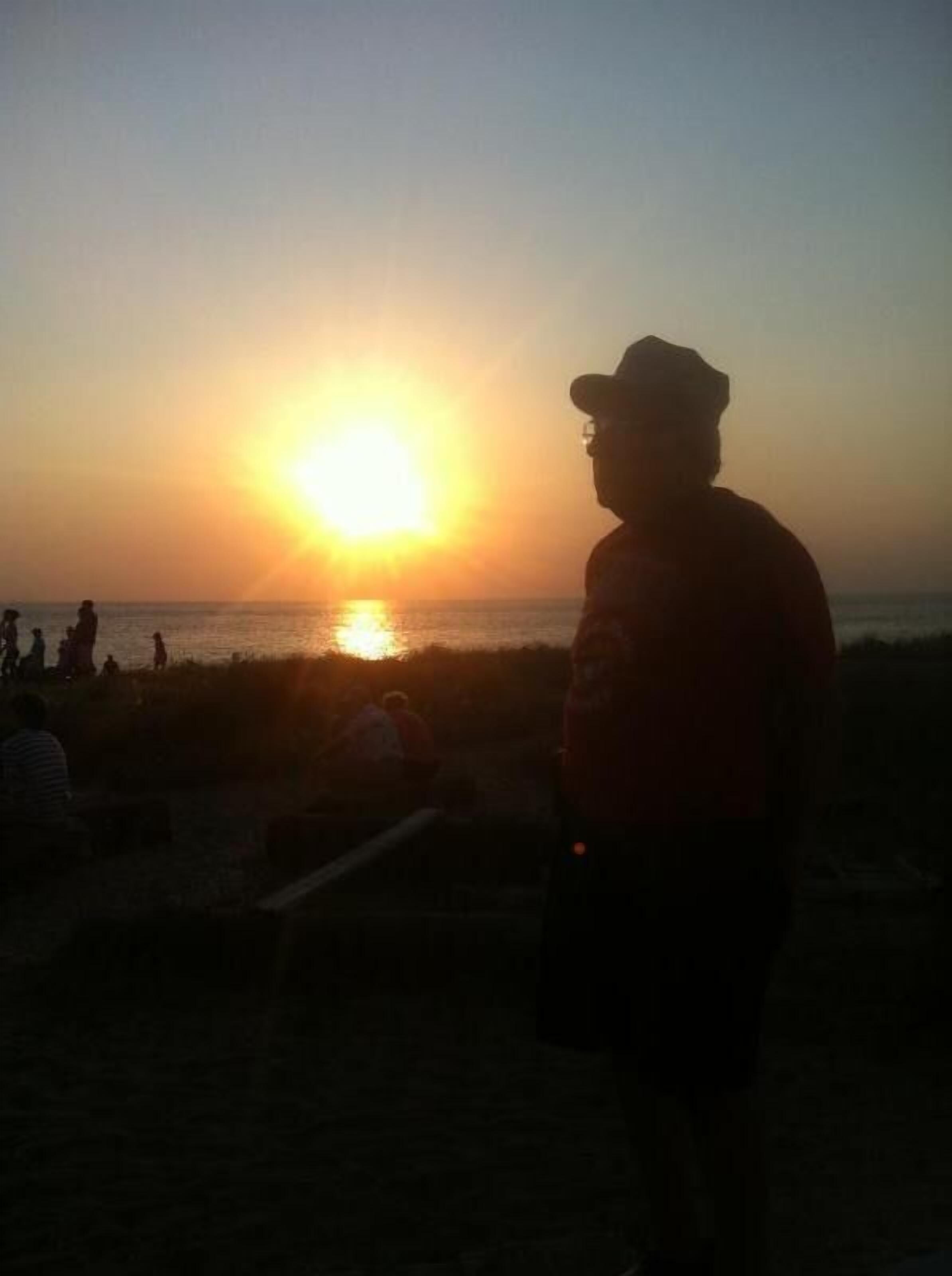 A beach scene at sunset with silhouettes of people and a person in a hat standing on the sand.