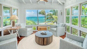 A spacious room with a large window offering a view of the ocean and palm trees, a round wooden coffee table, and two grey armchairs. - Luxury Beachfront Home in Haena AC, Pool  TVNC5141 (Hanalei)