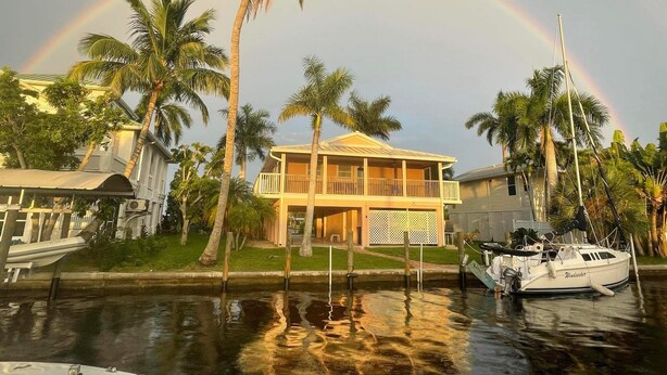 A waterfront property with a two-story house, a boat docked nearby, and a rainbow in the background. - The Bokeelia Boathouse, waterfront, Boat Dock, Pine Island, Florida (Bokeelia)