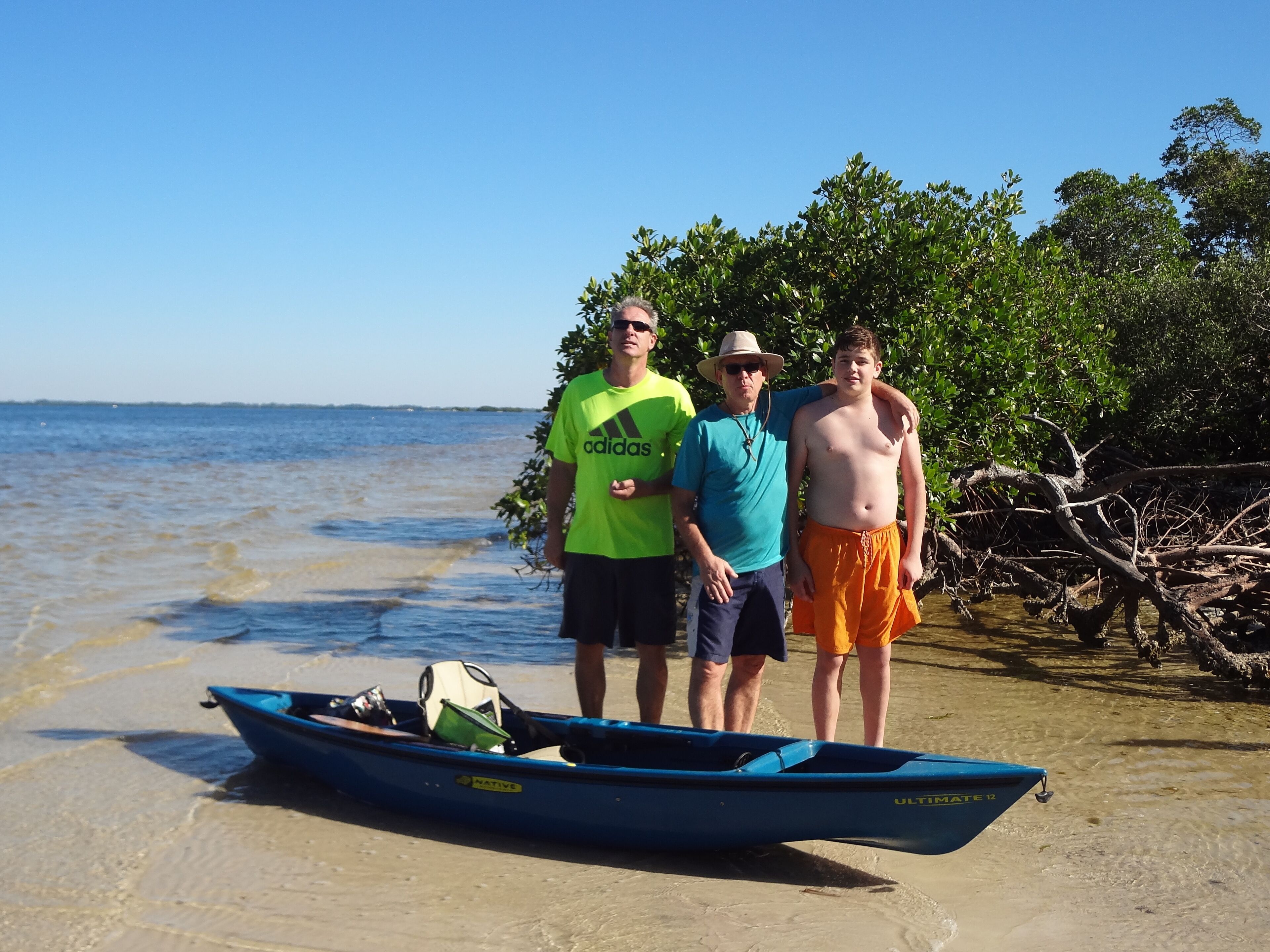 Trois personnes se tiennent près d’un bateau sur une plage, avec une eau claire et des mangroves en arrière-plan.