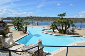 A swimming pool with stone steps and two stone lion statues, surrounded by palm trees and chairs, with a view of a lake and hills in the background. - Amazing top floor view in a newly renovated Lake Travis Condo on Private Island (Lago Vista)