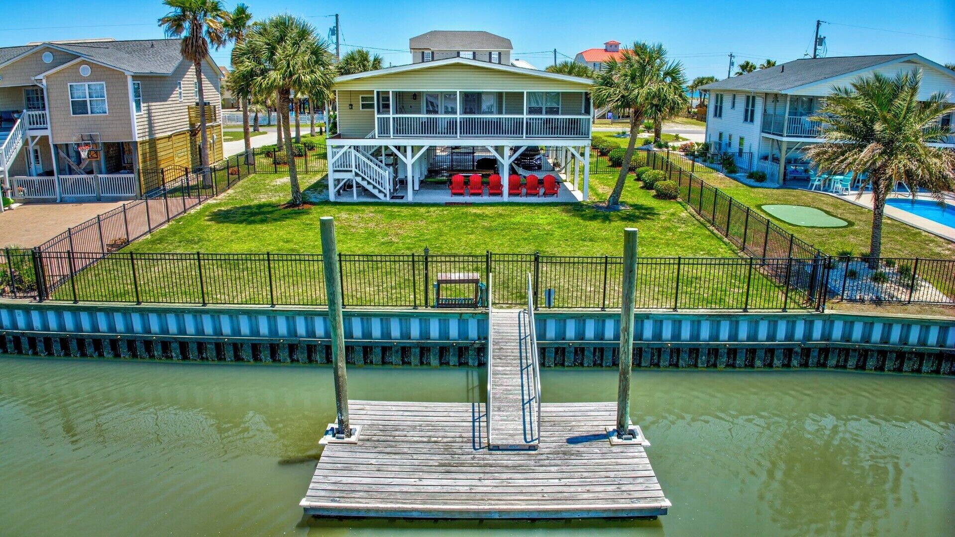 A dock leading to a house with a porch and red chairs, surrounded by a fence and palm trees.