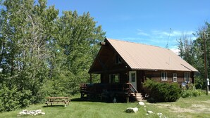 A rustic log cabin with a porch, surrounded by lush greenery and trees.