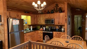 A wooden cabin kitchen with a stainless steel refrigerator, microwave, and stove. There is a dining table with chairs and a chandelier above. - Clemson Football- Lake Hartwell Lakefront Cabin Private Dock 4 BR (Fair Play)