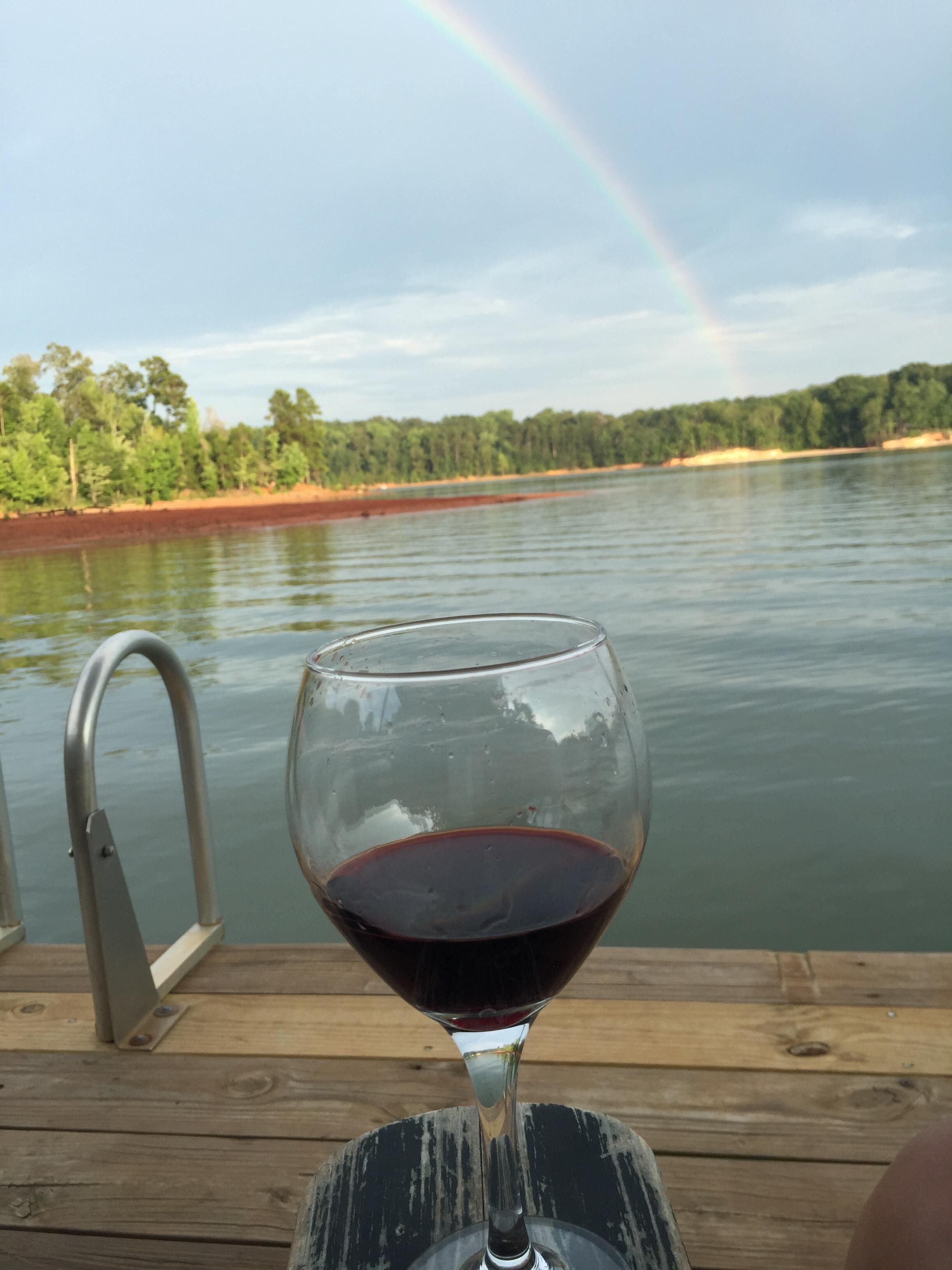 A glass of red wine on a wooden deck with a metal railing, overlooking a body of water and a forested shoreline.