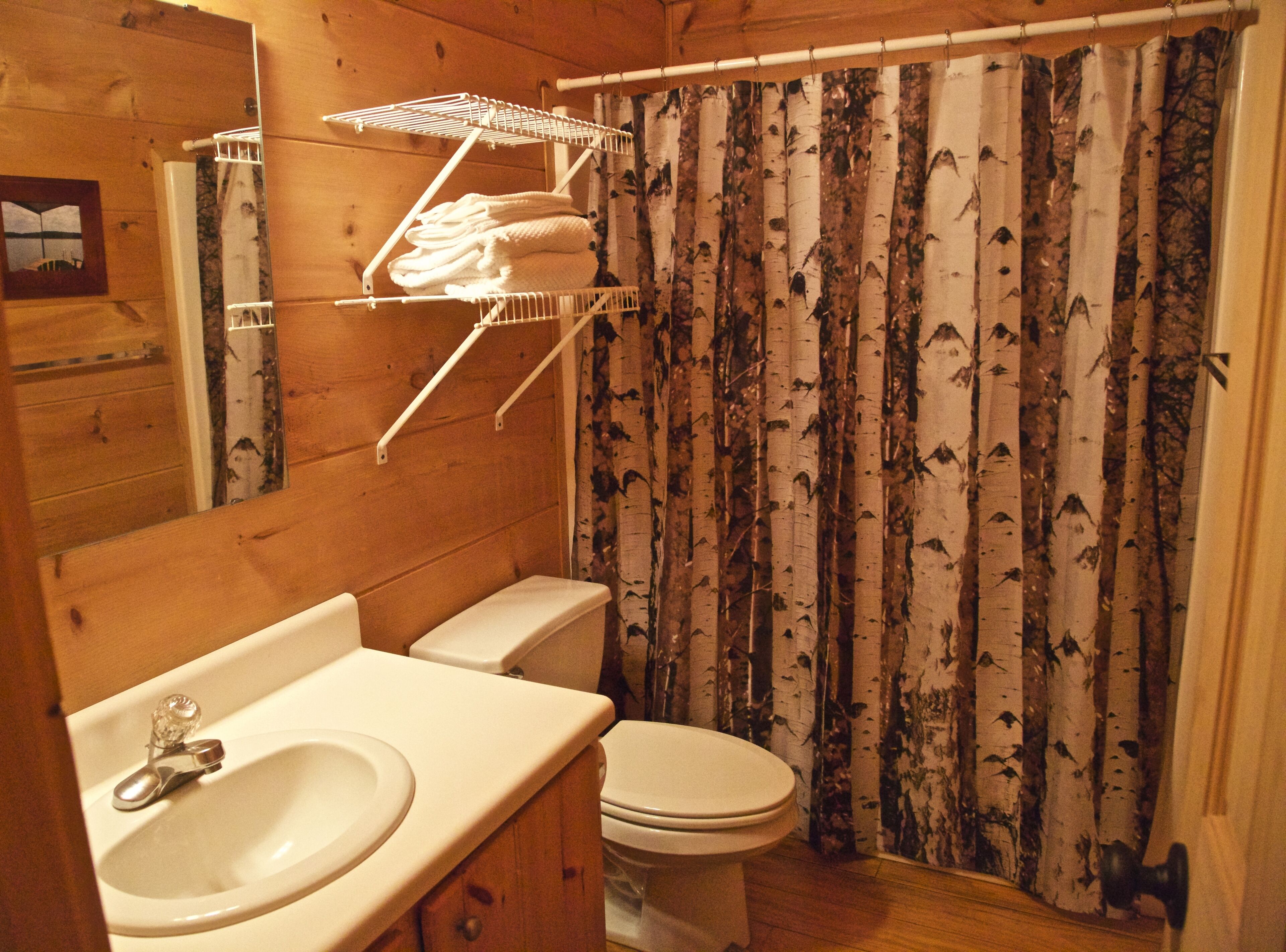 Baño de cabaña de madera con inodoro, lavamanos y repisa sobre el inodoro con toallas.