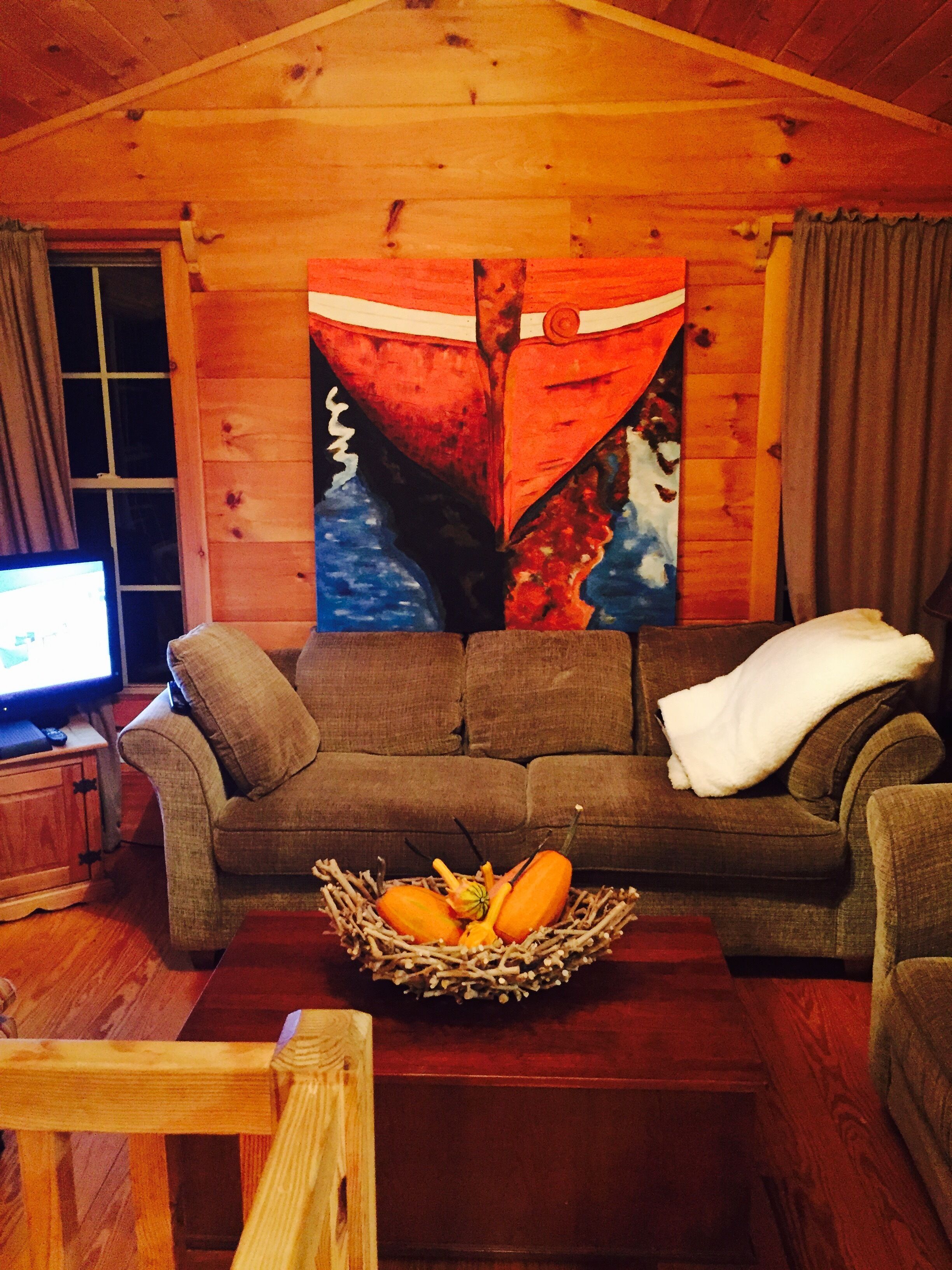 Ein gemütliches Blockhäuschen-Interieur mit Holzbalkendecke, einem braunen Sofa mit weißer Decke, einem Holztisch mit Obstschale und einem großen abstrakten Gemälde an der Wand.