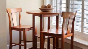A dining area with a round wooden table and two chairs, a framed picture on the wall, and a window with blinds.