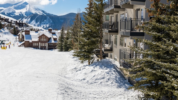 A snow-covered ski resort with a mountain in the background, a wooden lodge, and a modern building with balconies.