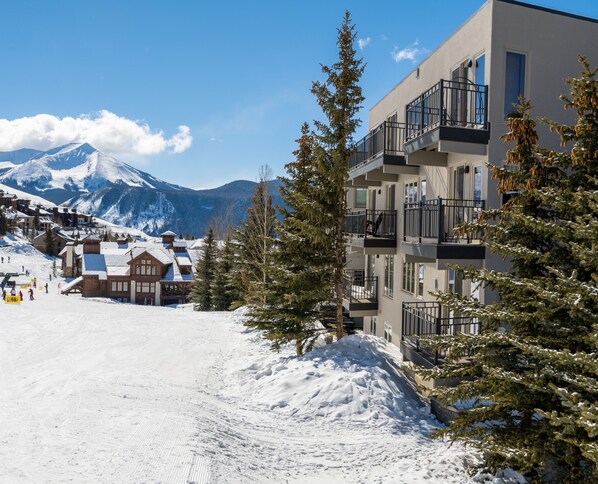 A snow-covered ski resort with a mountain in the background, a wooden lodge, and a modern building with balconies.