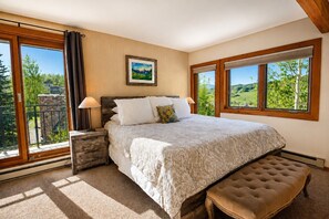 A bedroom with a large bed, two bedside tables, a bench, and a view of greenery through the windows. - True Ski In / Out with Amazing Views, Garage, Elevator & Fireplace (Crested Butte)
