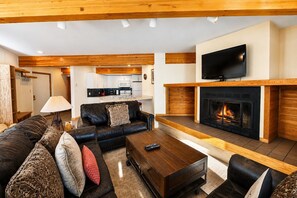 A living room with a fireplace, a television mounted above it, and a kitchen in the background. - True Ski In / Out with Amazing Views, Garage, Elevator & Fireplace (Crested Butte)