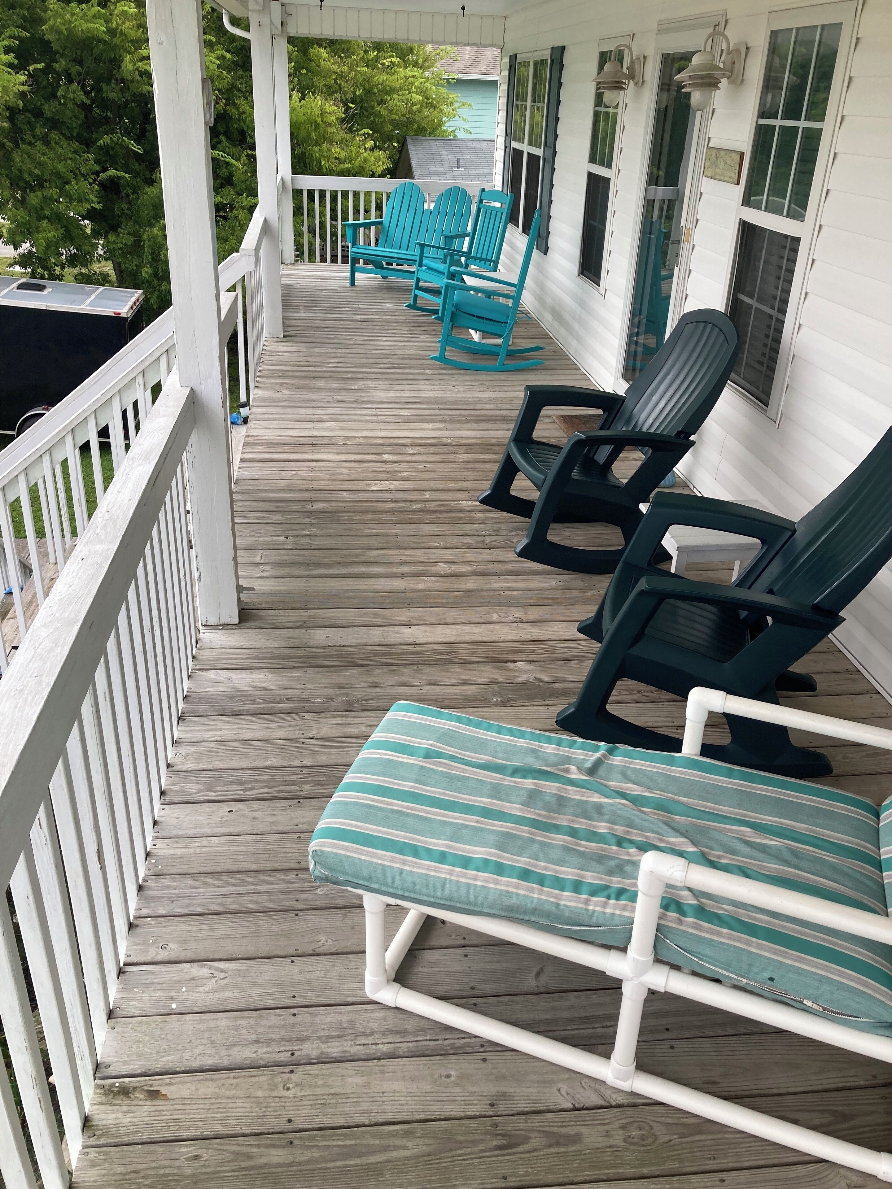 A covered porch with wooden decking and white railings. There are several lounge chairs, some with cushions.