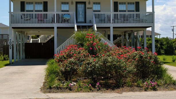 Ein zweistöckiges Haus mit einem Balkon im oberen Geschoss, einer Haustür und einem kleinen Garten mit rosa Blumen.