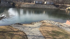 Un lago tranquilo con orilla rocosa, rodeado de árboles sin hojas y algunos edificios al fondo.