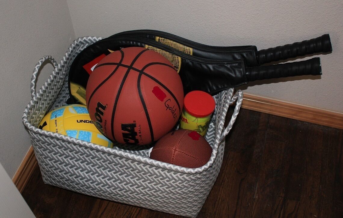 A woven basket containing sports equipment including a basketball, a football, and a tennis racket.