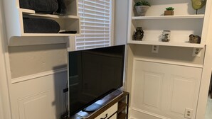 Un salon moderne avec un téléviseur à écran plat posé sur un meuble en bois, des étagères blanches intégrées ornées d’objets décoratifs et une fenêtre avec des stores.