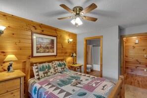 Une pièce chaleureuse aux murs en bois, avec un lit recouvert d’une courtepointe à motifs, un ventilateur de plafond et un tableau accroché au mur.