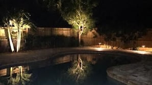 Vista nocturna de una piscina con borde curvo, rodeada por un patio de piedra y una valla de madera.