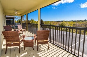 A balcony with wicker furniture, a ceiling fan, and a view of a parking lot and trees.