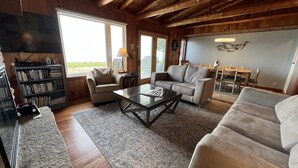 Ein gemütliches Wohnzimmer mit Holzbalkendecke, einem Bücherregal, zwei Sofas, einem Couchtisch und einem Essbereich mit Tisch und Stühlen.