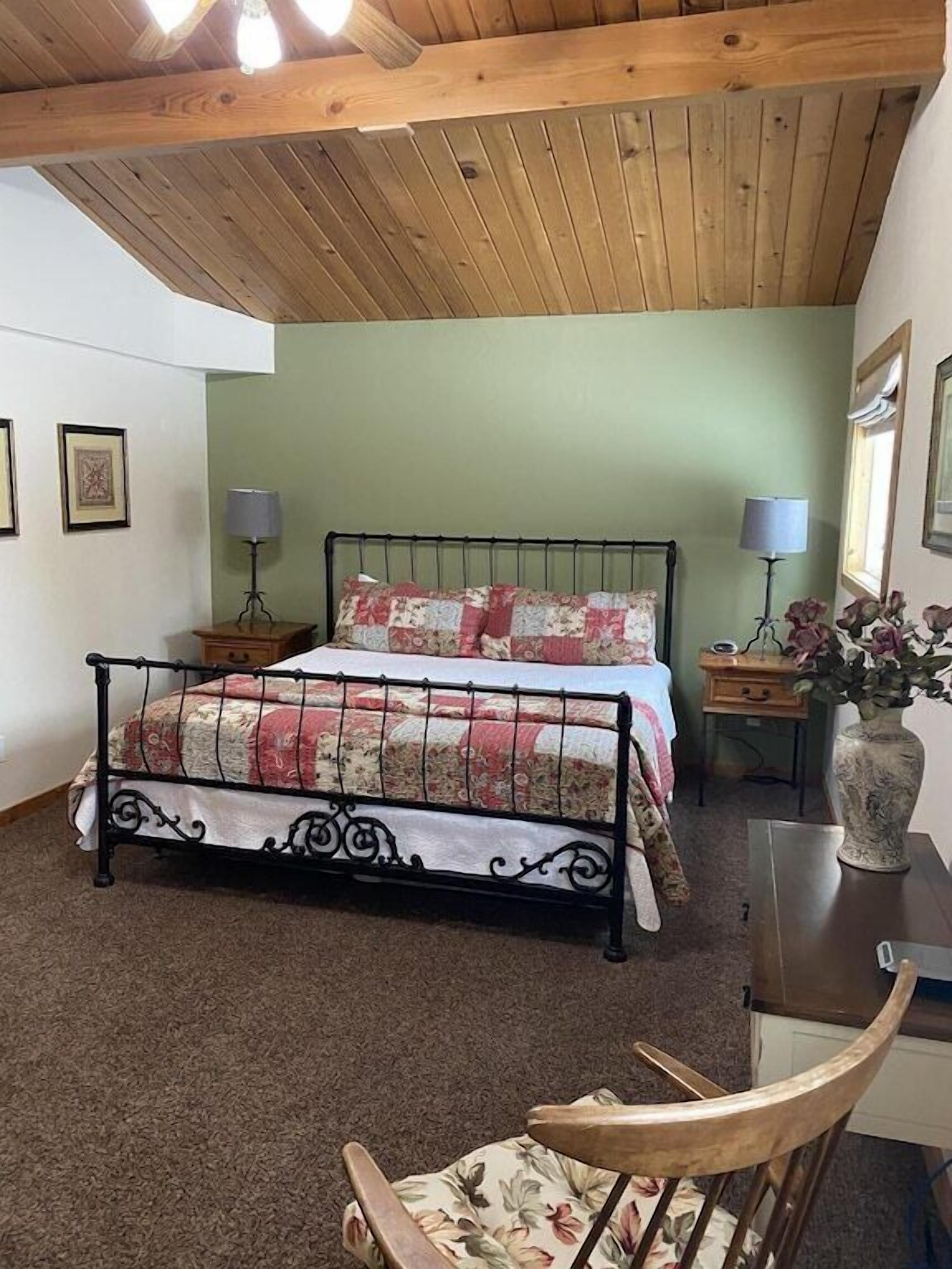Un dormitorio con estructura de cama metálica, colcha de estampado floral, una silla de madera y un escritorio con un florero.