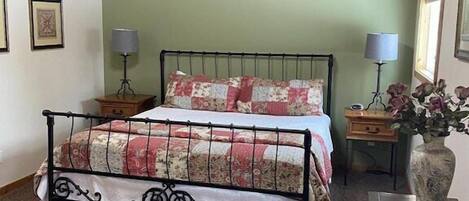 Un dormitorio con estructura de cama metálica, colcha de estampado floral, una silla de madera y un escritorio con un florero.
