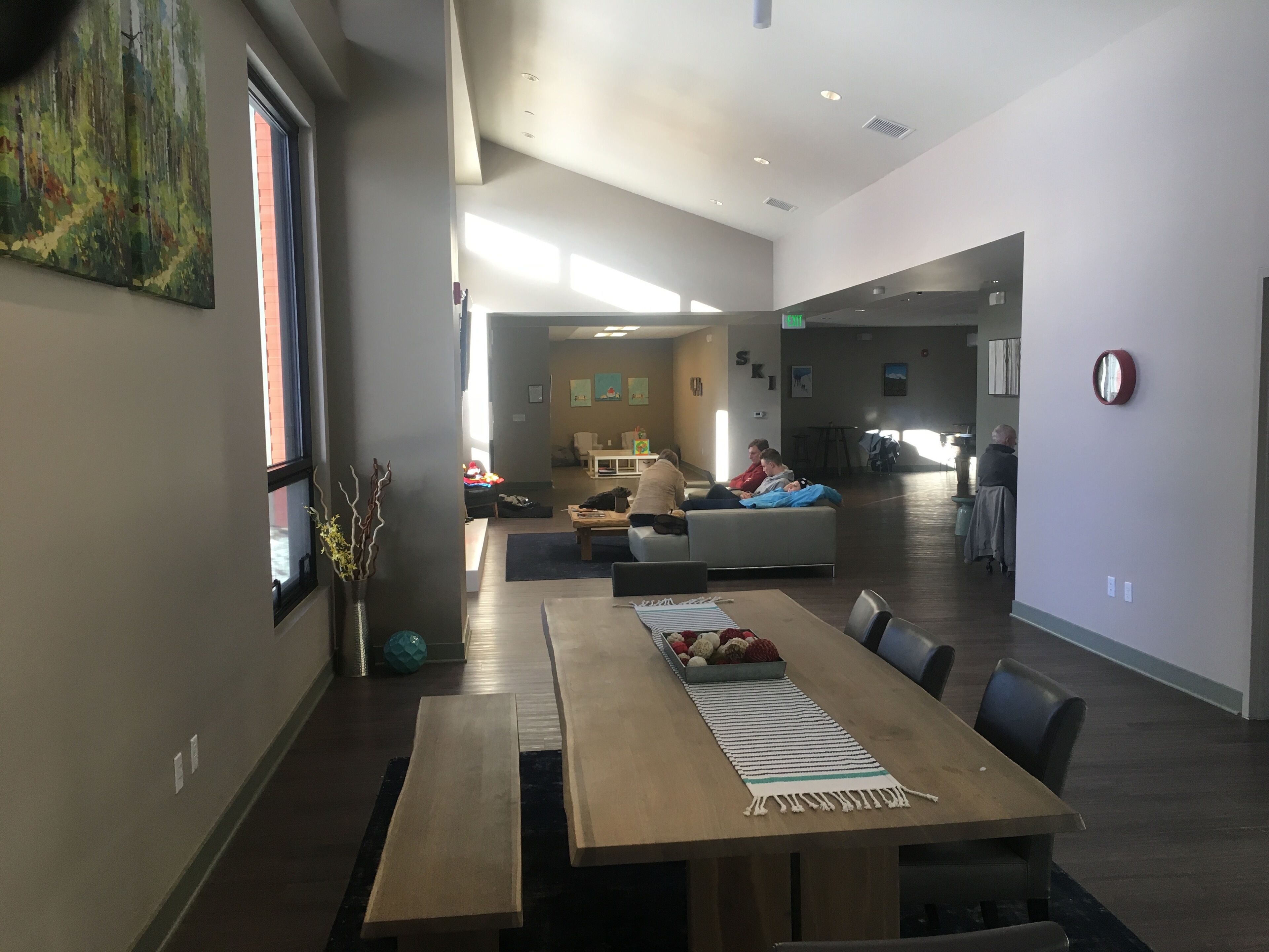 A spacious indoor area with a long wooden table, several chairs, and a couch. There is a large window on the left, a painting on the wall, and a clock on the right.