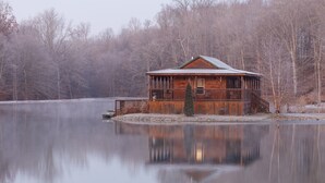 Une cabane en bois, surmontée d’un toit en métal, située au bord d’un lac paisible et entourée d’arbres.