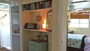 A compact room with a bed, a small desk with a lamp, a bookshelf, and a white chair. -  Cute Cozy Cottage with private entrance and ocean views in prime neighborhood (St. John)