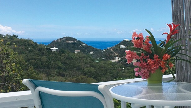 A balcony with a table and chairs overlooking a coastal landscape with houses and a view of the sea. -  Cute Cozy Cottage with private entrance and ocean views in prime neighborhood (St. John)