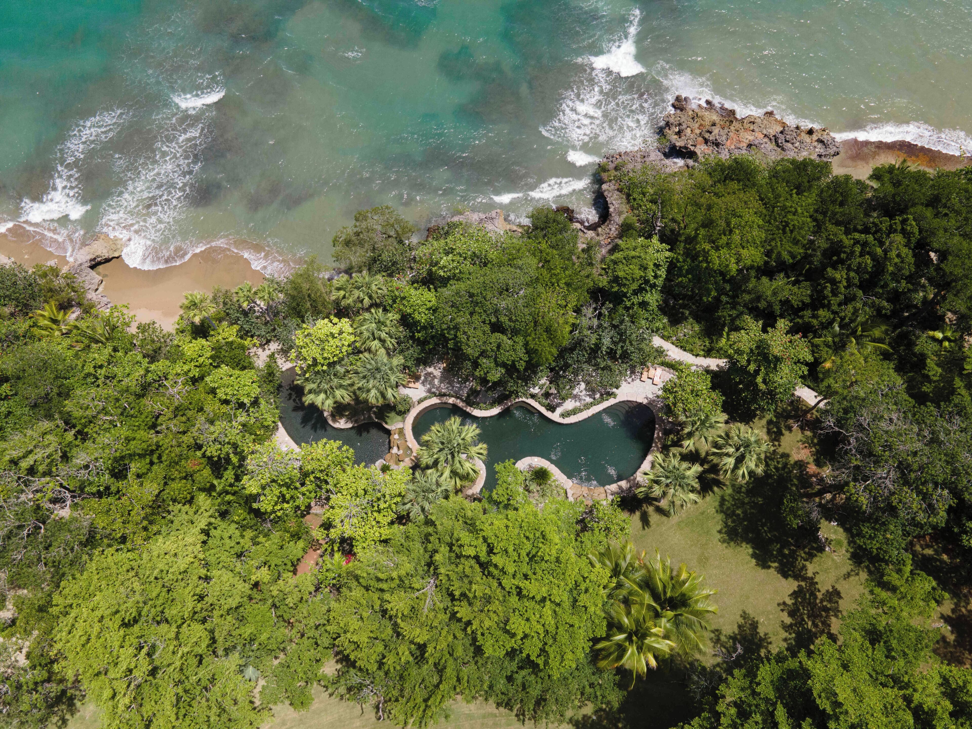 An aerial view of a tropical resort with a central swimming pool surrounded by dense greenery and a rocky coastline.