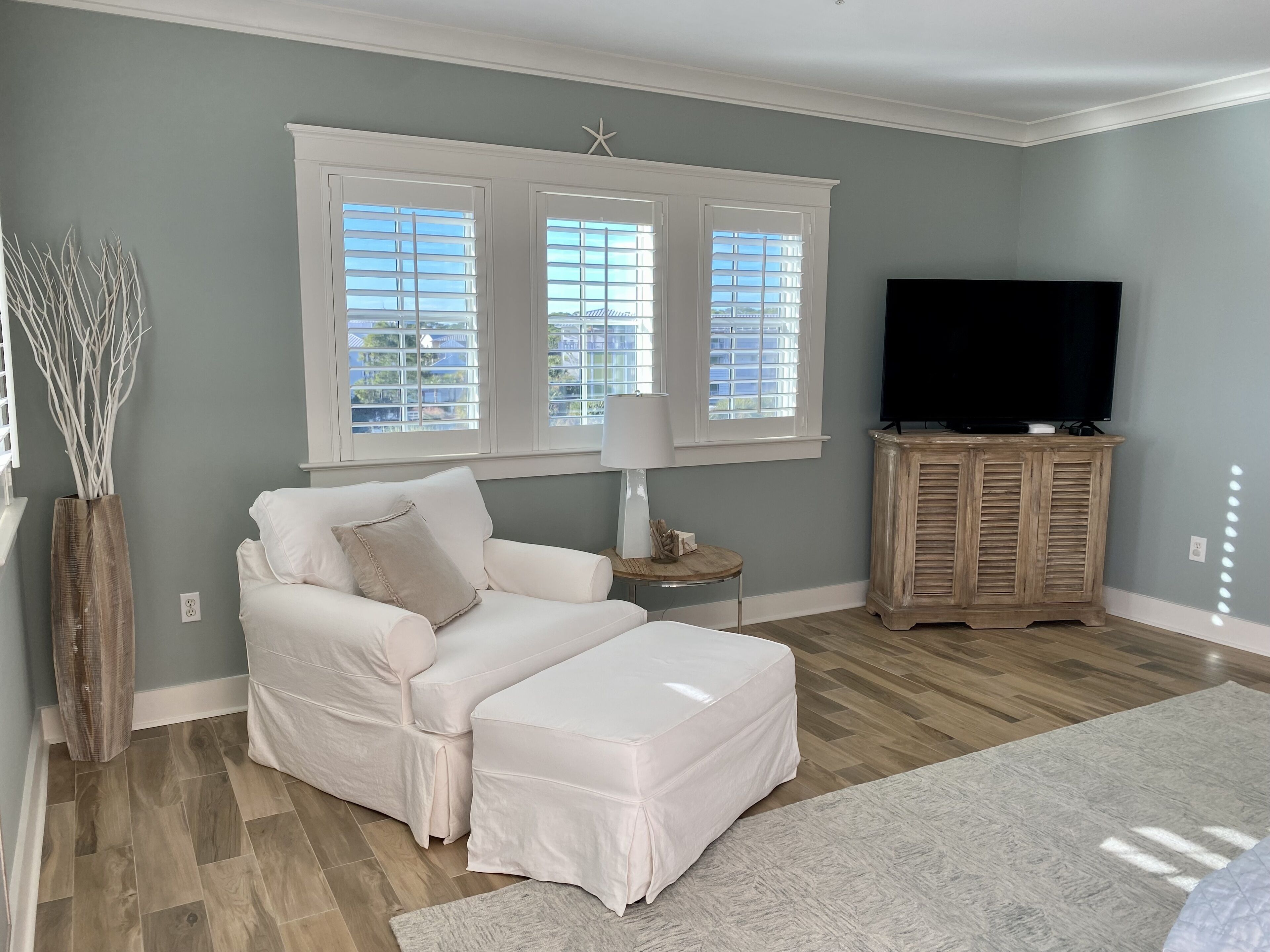 A living room with a white sofa, ottoman, and a TV on a wooden cabinet. There are three windows with blinds, a lamp on a side table, and a decorative starfish on the wall.