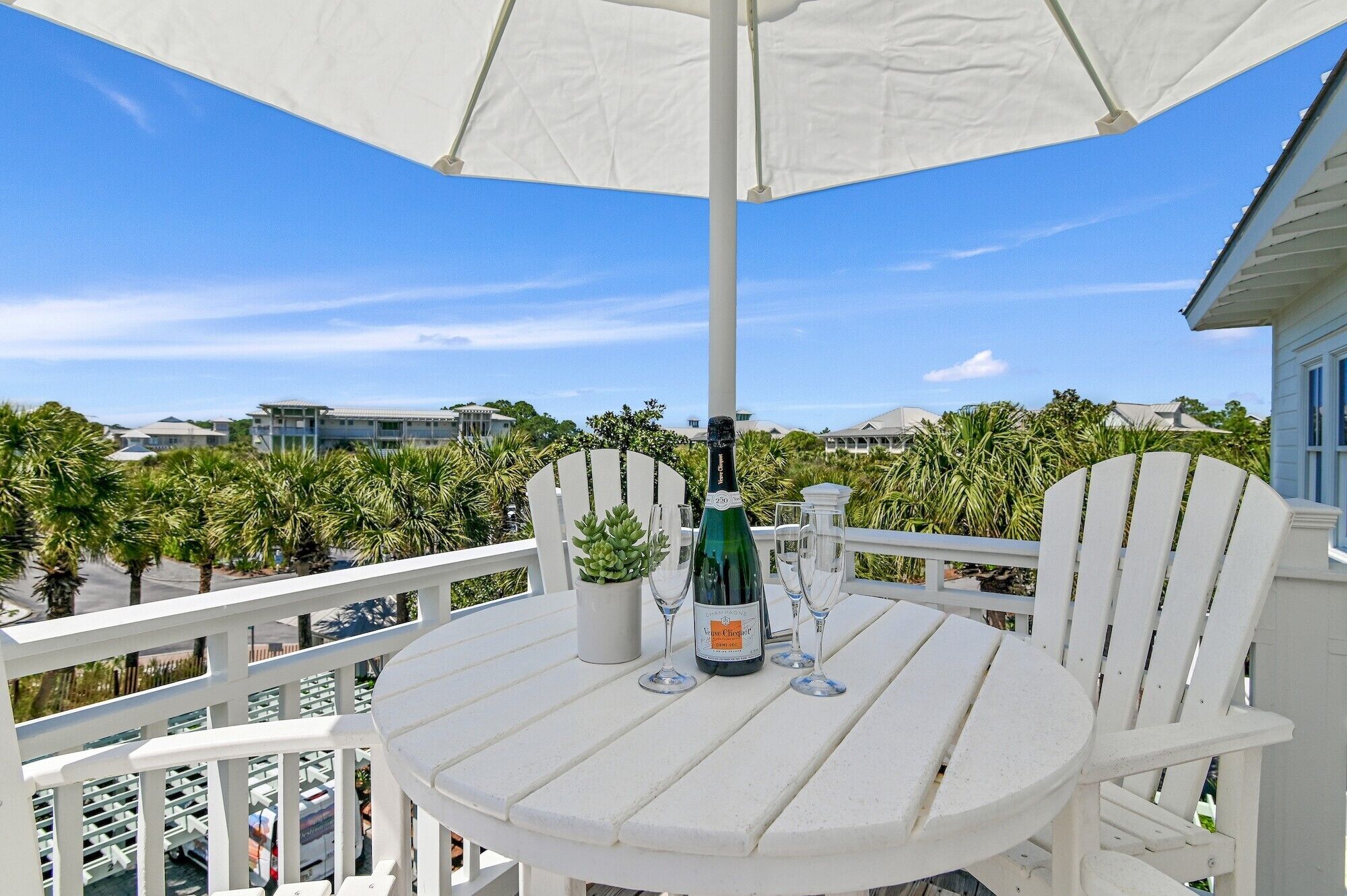 A white outdoor table with a bottle of champagne and two glasses, surrounded by white chairs, under a white umbrella, with a view of palm trees and buildings in the background.