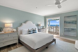 Une chambre à coucher avec un lit, des tables de chevet, un ventilateur de plafond et une vue sur l’océan par une porte ouverte.