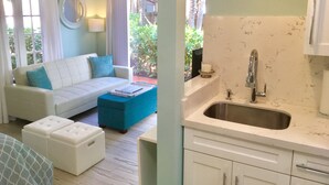 Un salon moderne avec un canapé blanc, des coussins bleus et un pouf turquoise. On aperçoit un coin cuisine avec un évier et des meubles blancs.