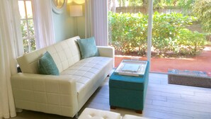 Un salon avec un canapé blanc, un pouf turquoise et une table basse en verre.