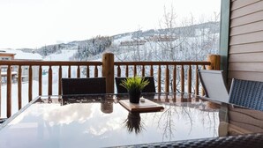 A glass-top table with a small plant on a wooden coaster, a chair, and a view of a snowy landscape through a wooden railing.