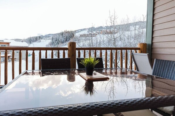 A glass-top table with a small plant on a wooden coaster, a chair, and a view of a snowy landscape through a wooden railing. - KR410 by Simply Kimberley | Ski-In/Out + Private Hot Tub (Kimberley)