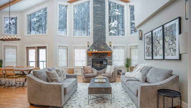 A spacious living room with a stone fireplace, large windows with snowy views, and a high ceiling.