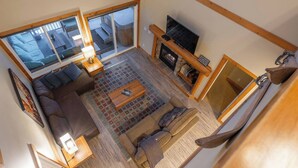 A spacious living room with a large sofa, a coffee table, and a fireplace. There is a television on a wooden stand and a large window providing natural light.