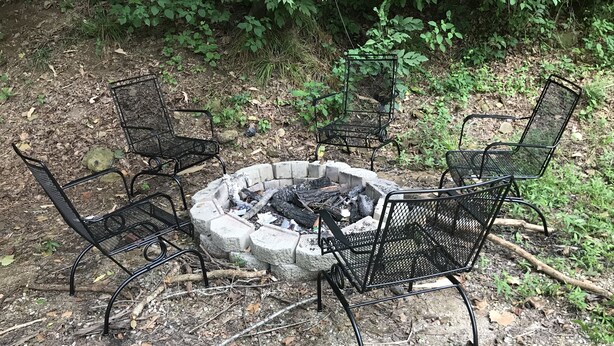 A fire pit with four metal chairs arranged around it in a forested area.
