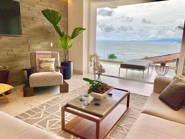 A modern living room with a large window overlooking the sea, a comfortable sofa, a coffee table, and a potted plant.
