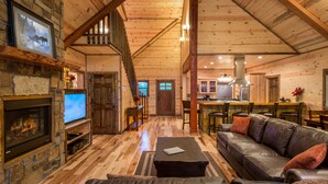 A spacious living room with a fireplace, a stone fireplace, a wooden staircase, a kitchen area, and a dining table. - Crystal Ridge, Wet Spa, Pool Table, Arcade Game, Hot Tub, Wood-burning Fireplace (Broken Bow)