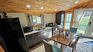 A compact kitchen with a black refrigerator, wooden cabinets, and a dining area with a table and chairs. - The Byre Bungalow--Country Getaway, close to Yough, Fallingwater w/ Hot Tub! (Confluence)