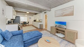 A living room with a blue sofa, a round coffee table, a television, and a kitchen area in the background. - Pelican Perch: Tropical Paradise Cottage on the Beach            VRC17-000167 (Holmes Beach)