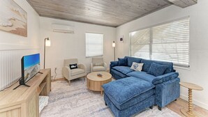 A living room with a blue sectional sofa, a round coffee table, a TV on a wooden stand, and a patterned rug. - Pelican Perch: Tropical Paradise Cottage on the Beach            VRC17-000167 (Holmes Beach)