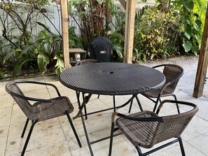 Une table ronde d’extérieur, entourée de quatre chaises en osier, le tout agrémenté d’une végétation luxuriante et d’une pergola en bois.