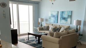 A spacious living room with a beige sofa, a coffee table, and a dining table with chairs. There is a ceiling fan, two wall-mounted lamps, and a large window with a view of the outdoors. - Dog Days of Summer on the Beach! August/Fall Dates Open
Cyndi 386-361-5882 (Daytona Beach Shores)
