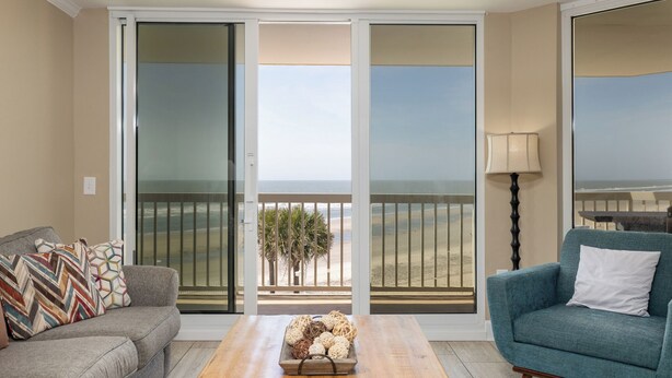 Ein Wohnzimmer mit einer Couch, einem blauen Sessel, einem Couchtisch aus Holz und Blick auf den Strand durch eine Schiebetür.